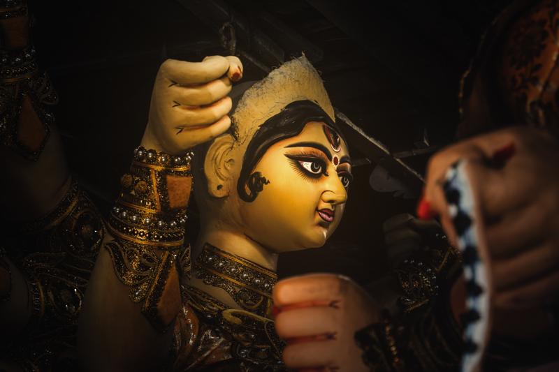 A brush approaches the idol’s eye in close view, revealing the fine gold line, dark lashes and warm clay beneath the pigment.