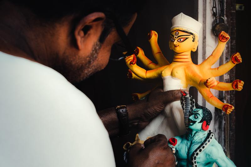 A man steadies a small statue while working in the crowded workshop, his blue clothing bright among the surrounding unfinished idols.