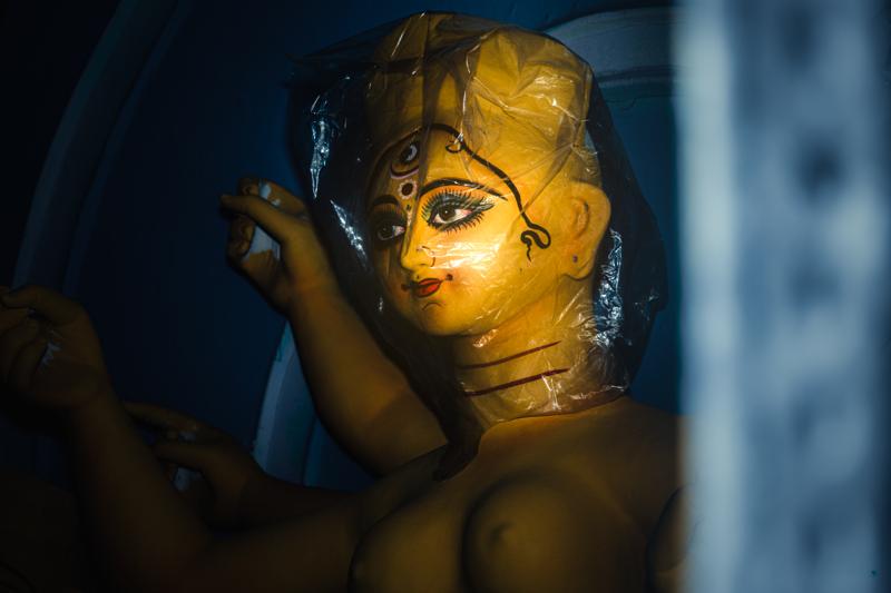 One painted face emerges from the shadow, its wide eye and gold details surrounded by the blurred shapes of the crowded workshop.