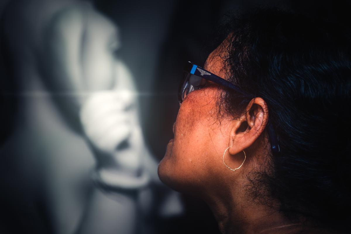 A woman’s profile fills the edge while an idol’s painted eyes face forward behind her, joining observer and figure in one frame.