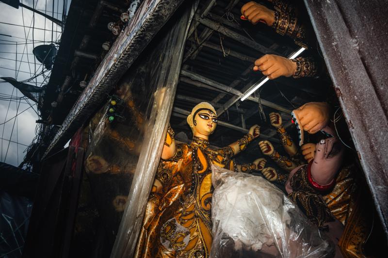 A painted goddess appears through a narrow workshop opening, her golden face and raised hand catching light among beams and unfinished forms.