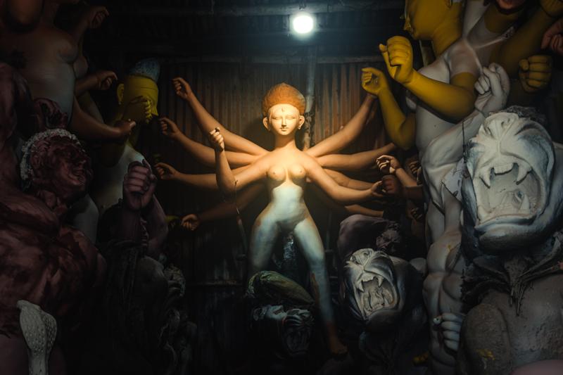 The central clay figure glows beneath a bare bulb, while rows of other forms recede into the packed, shadowed workshop.