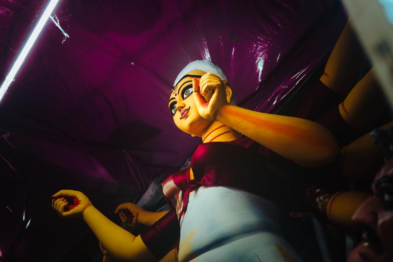 A bright painted face turns through the purple-lit workshop, its yellow features and lifted hand isolated against the dark surroundings.