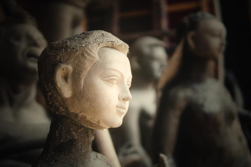 A pale unfinished head fills the foreground while the artisan’s face appears beyond it, both profiles held in the workshop’s warm light.