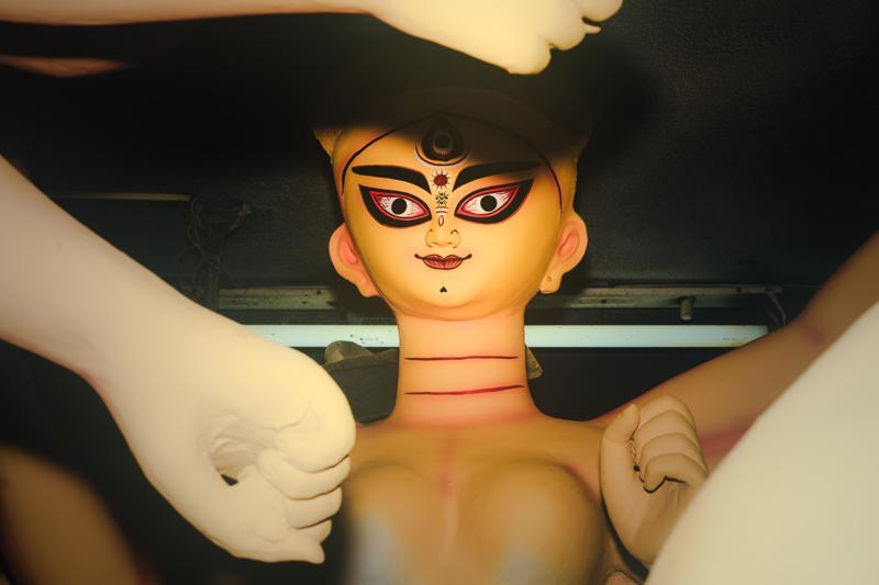 The clay figure’s painted face and many arms fill the close frame, with hands and yellow light separating it from the room.