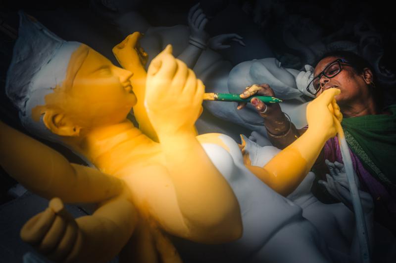 A focused worker reaches toward the goddess’s face, his hand and brush catching the lamp’s glow beside the smooth yellow clay.