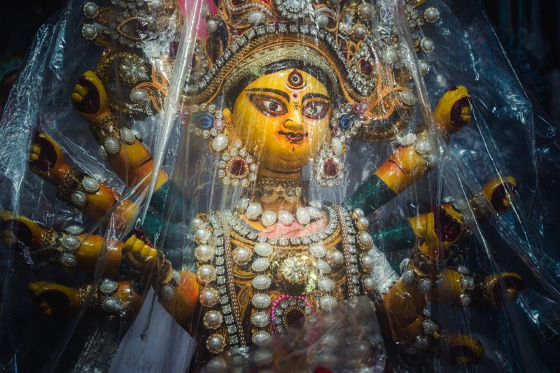 The goddess’s gold face emerges through reflective decoration; tiny beads and dark folds gather around her wide eyes beside an ornate crown.