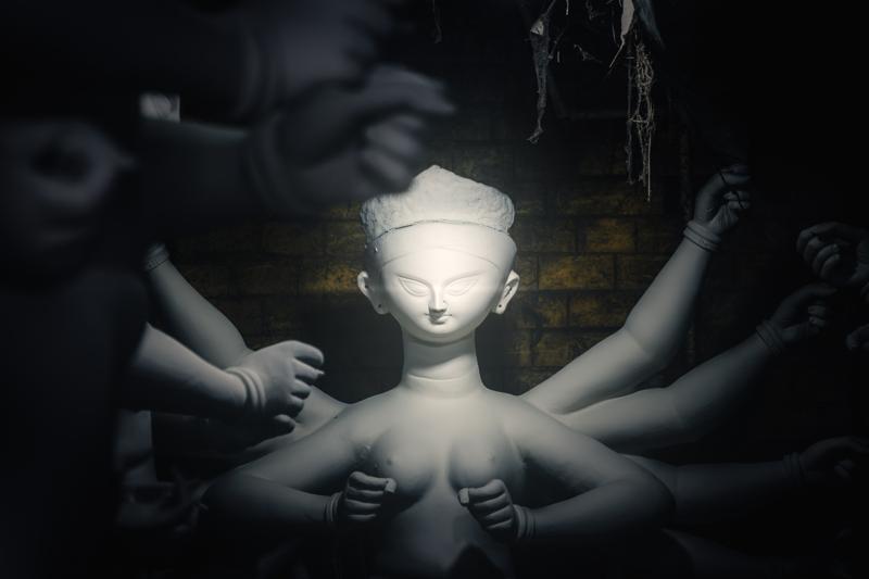 The idol’s sculpted face appears under a cool pool of light, its pale forehead and raised hands surrounded by deep blue shadow.