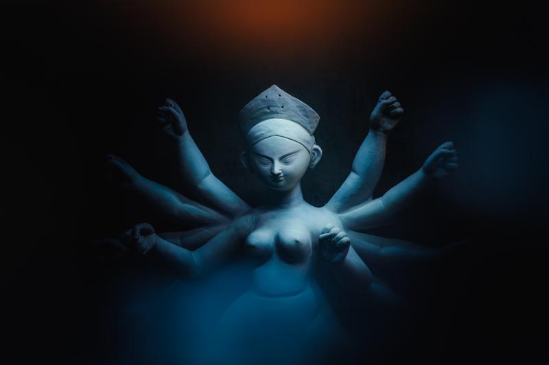 A focused worker bends beside a soft clay figure in the packed workshop, the pale statues pressing close around his bare shoulders.
