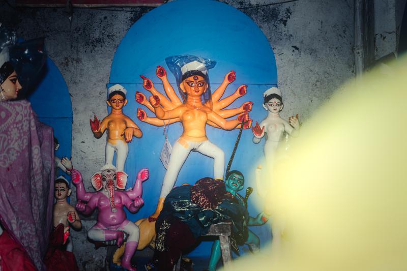 A painted Durga stands with attendants beneath a blue circular backdrop, while yellow blur obscures part of the shrine’s lower right foreground.