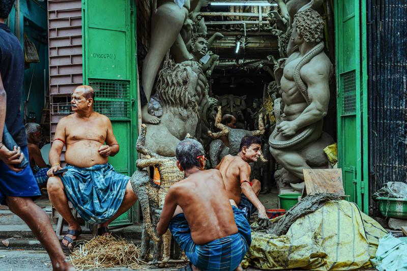 A sculptor bends close to a large clay figure, his bare back and the unfinished forms packed together in the dim room.