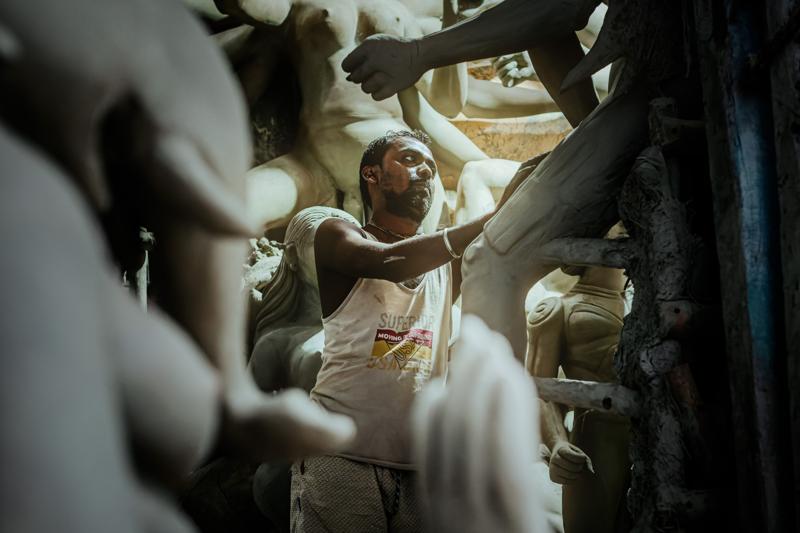 An artisan’s arm extends between pale clay bodies toward the brighter end of the workshop, his face partly hidden by the forms.