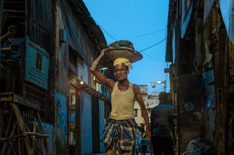A man walks toward the camera beneath a wide umbrella, his yellow vest and blue street walls bright in the midday sun.