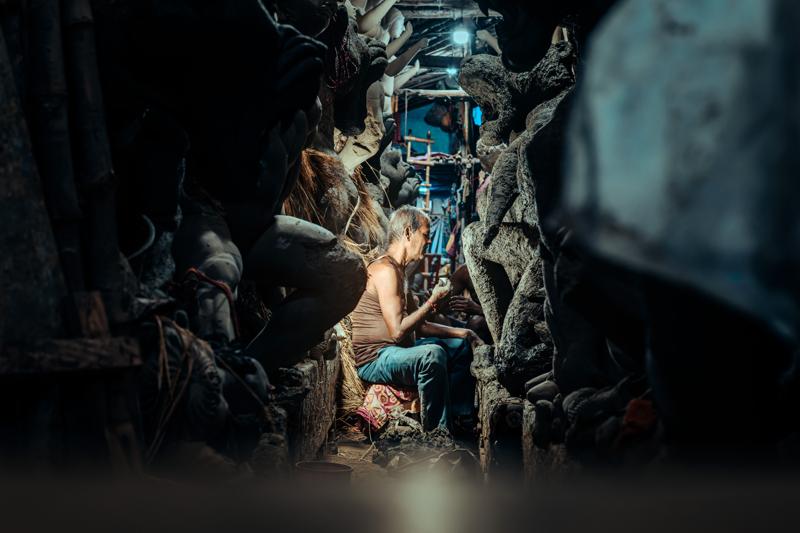 A worker kneels among unfinished figures in a dim corridor, where the small bright opening at the end gives the scene depth.