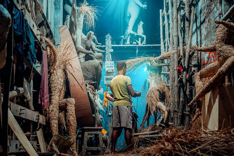 A focused worker moves toward the blue-lit opening between closely packed statues, his small figure anchoring the long workshop view.