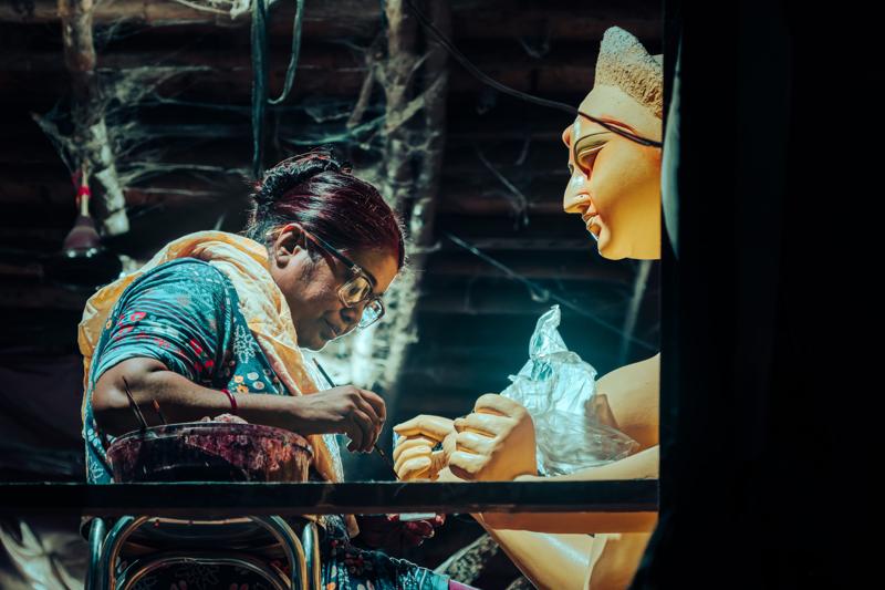A woman leans toward the bright clay face, her reflection joining the painted idol’s features in the marked glass of the stall.