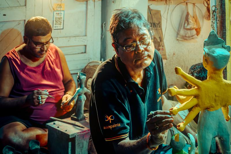 A woman and man sit near the bright yellow figure, their faces turned toward it within the busy workshop scene.