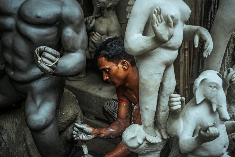 A sculptor works beside a large unfinished figure, surrounded by clay bodies and carved forms stacked through the cramped, dim workshop room.