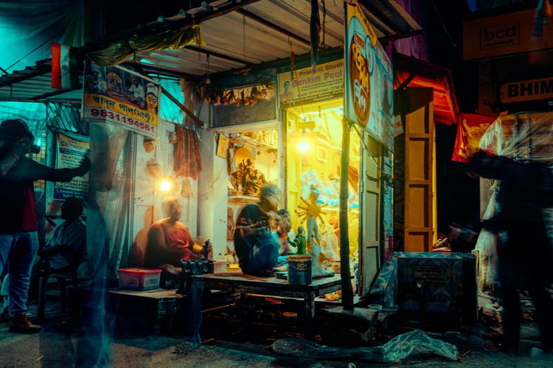 Warm light falls across the focused artisan and his clay figures, while the open doorway reveals the narrow market lane beyond.