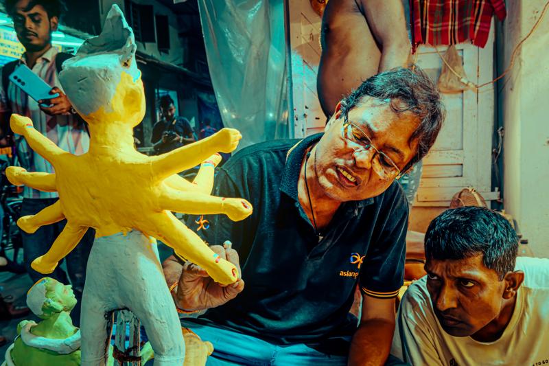 The seated sculptor leans toward a small yellow figure with an easy smile, while another worker looks on from the crowded shop.