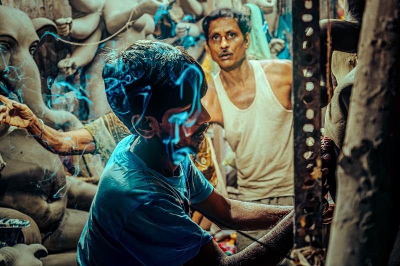 Two workers lean toward a clay figure in the busy studio, surrounded by unfinished statues and carved forms beneath the crowded roof.