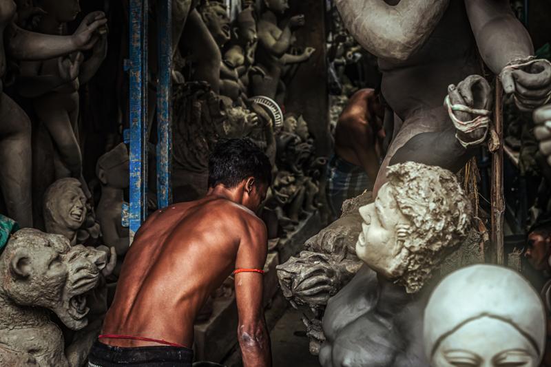 A shirtless artisan bends over a clay figure in the crowded workshop, while faces and unfinished forms surround him beneath the beams.