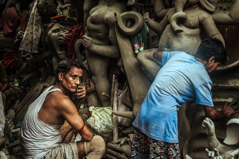 A focused worker stands between unfinished figures as light pours through the bamboo roof, revealing the crowded height of the crowded workshop.