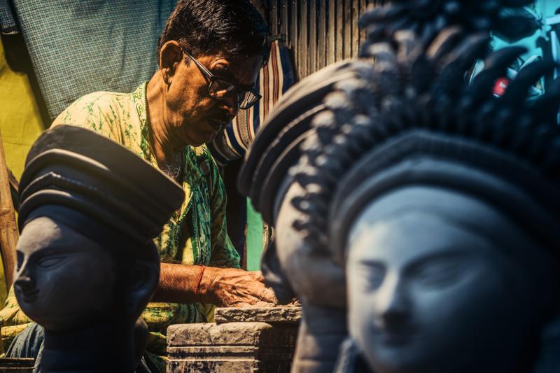 A man leans toward the idol, applying paint near its face as the large clay figure fills the foreground of the workshop.