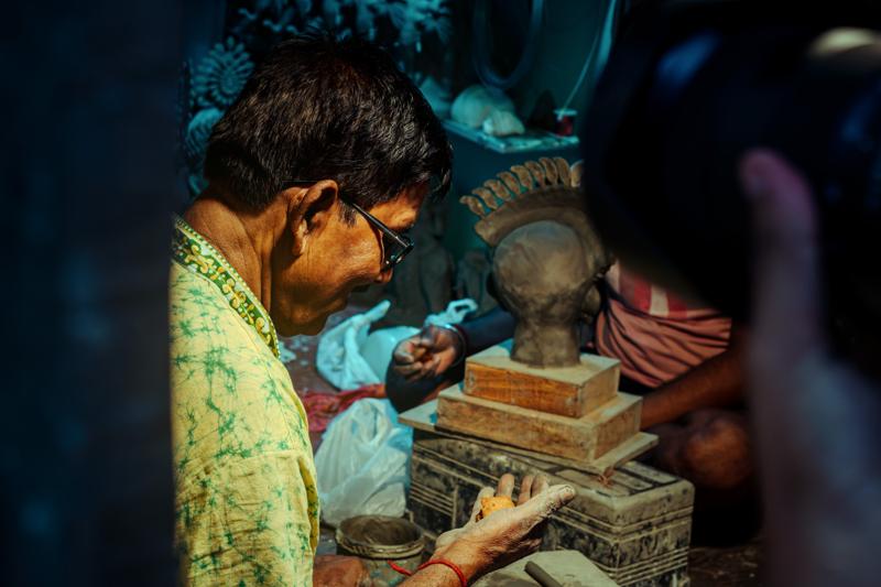 The artisan works close to a tiny clay head, his focused profile and the unfinished figure sharing the pool of workshop light.