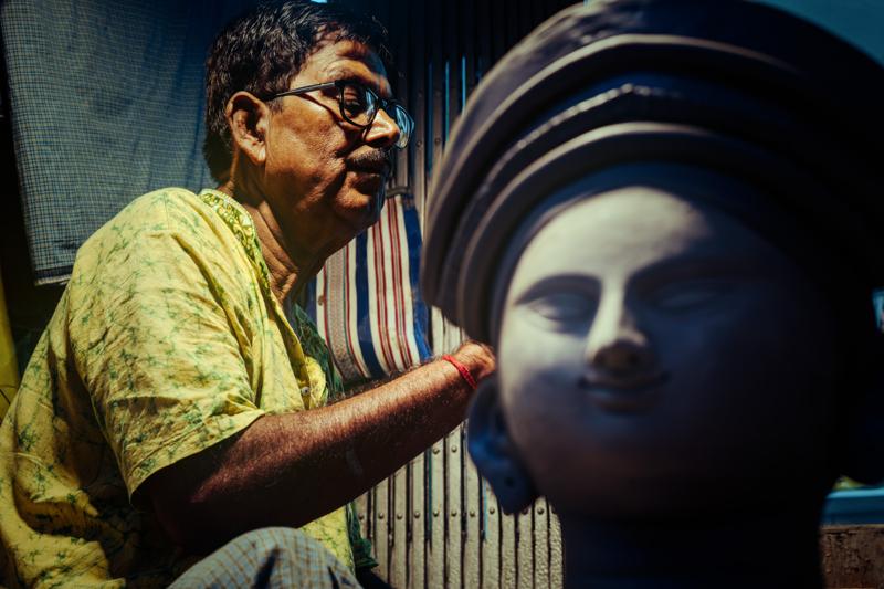 A bespectacled artisan leans toward the soft clay portrait, his cheek and the unfinished statue’s smooth profile catching the same warm light.
