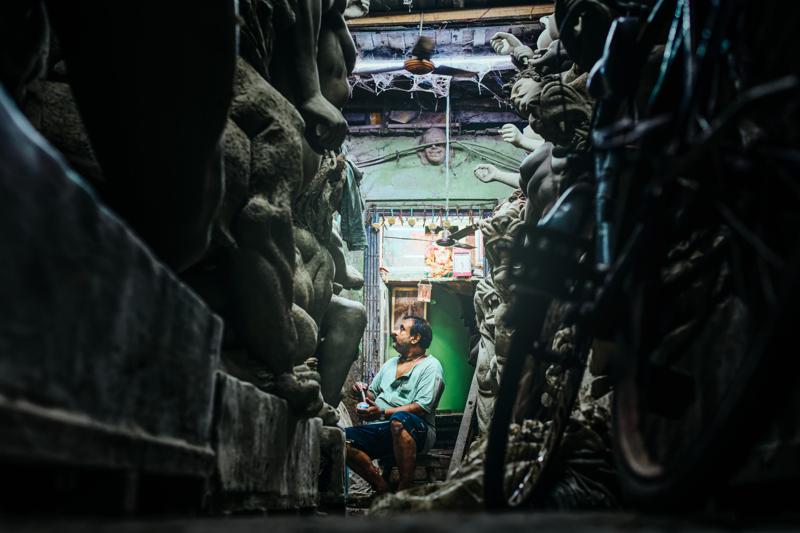 A seated figure appears at the end of the narrow lane, enclosed by stacked statues and rough walls on both sides.