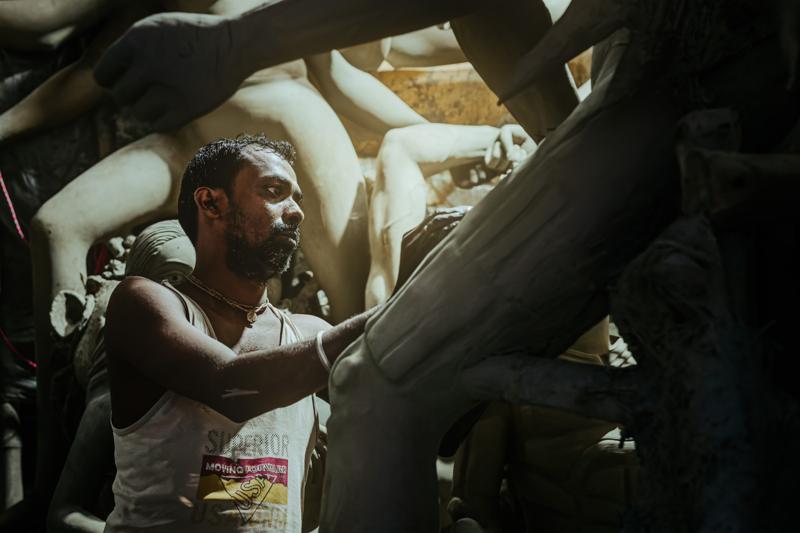A man reaches upward toward an unfinished figure, the bright opening above catching his raised arm amid the dark workshop forms.