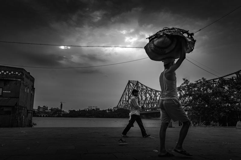 A man balances a broad load overhead as another figure crosses the riverbank; the steel bridge’s lattice fills the cloudy sky behind them.