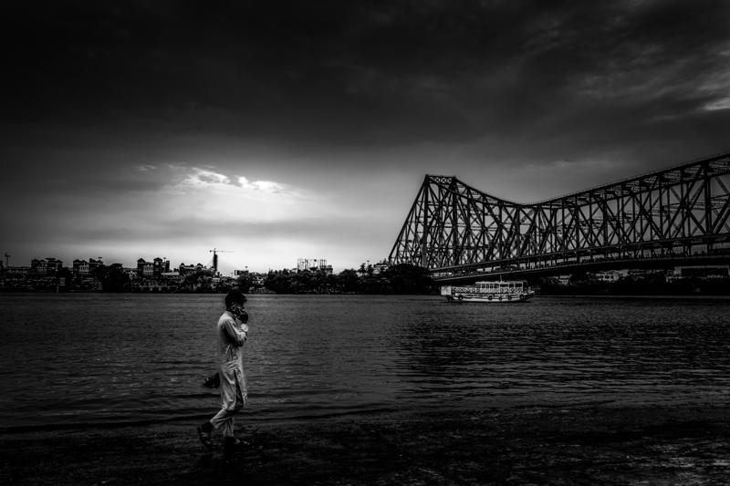 A lone woman walks beside the river beneath the immense bridge, her pale clothes set against dark water and the bright open sky.