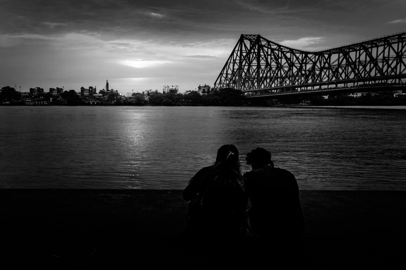 A pair sits facing the broad river beneath the steel bridge, their dark outlines small against the open monochrome water and low cloud.