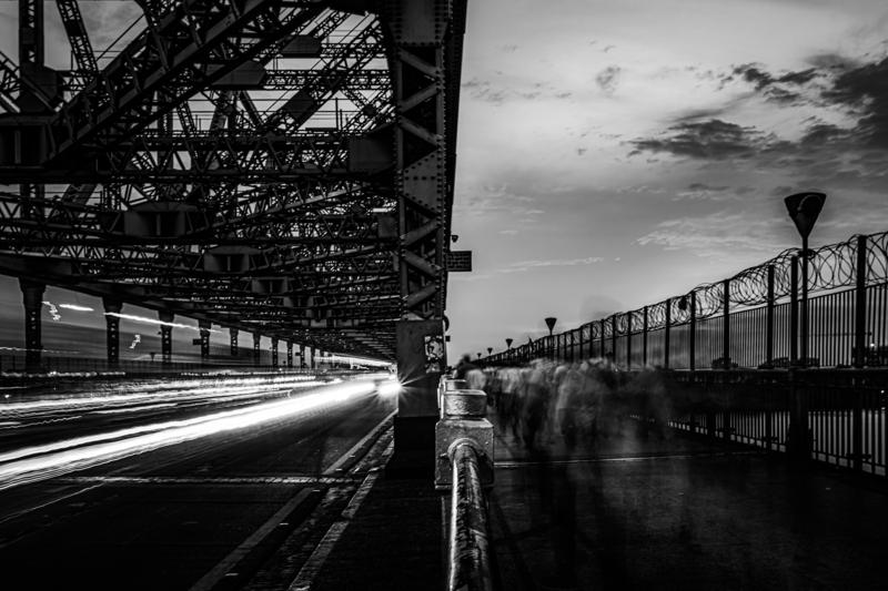 Two men sit by the dark river as the bridge spans the clouded sky; a track of water leads toward the distant bank.