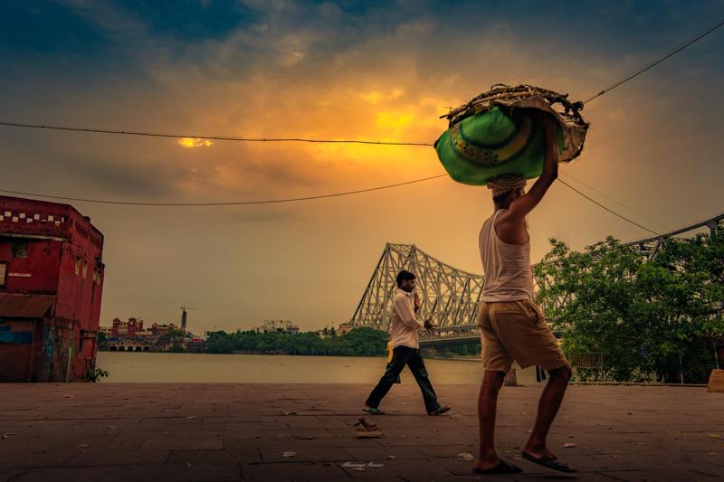 A porter steadies a rounded bundle overhead while walking beside the Hooghly; the steel bridge and amber clouds hold the bank in shadow.