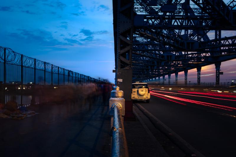Traffic becomes red and gold lines along the waterside road, while the steel bridge rises above the long-exposure glow, nearby, at the riverbank.