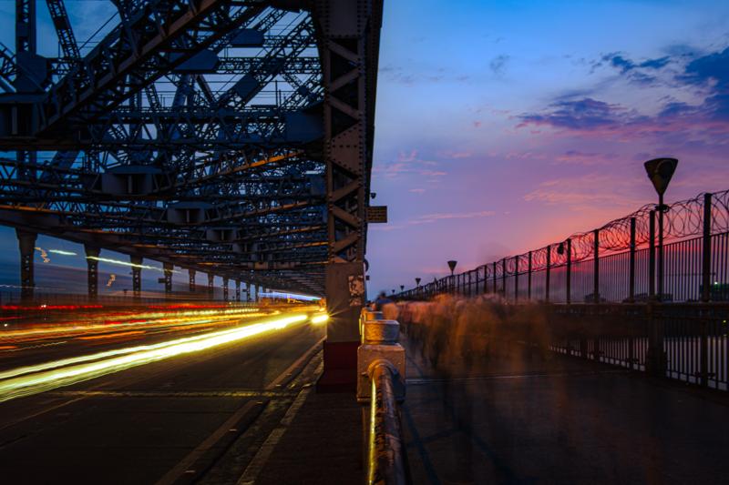 Steel beams fill the upper frame as traffic streaks across the broad river road, leaving the waterside lamps reflected below, nearby, over water.