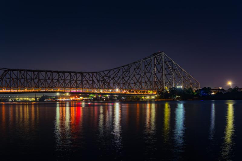 The illuminated bridge spans the night river, its warm lights repeated in long, broken strokes across the black water, nearby, under the bridge.