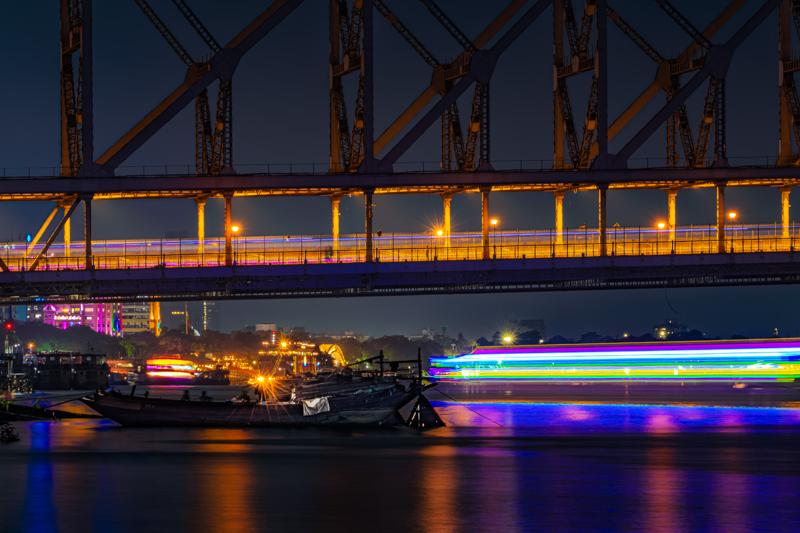 A small boat passes beneath the steel span as purple and amber lights shimmer across the still water after dark, nearby, at dusk.