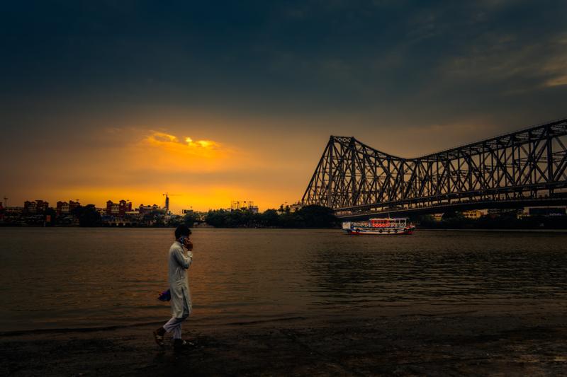 A lone man walks with a covered basket balanced overhead; the steel bridge spans behind him as the low sun warms the riverbank.