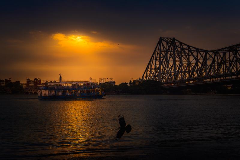 The steel bridge spans the amber river as a small figure steps through the shallows, isolated between the bright horizon and dark city.