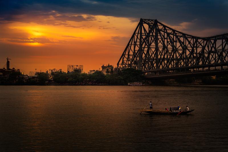 Orange light spreads across the water beneath Howrah Bridge, where a lone figure walks the bank and the far shore fades into dusk.