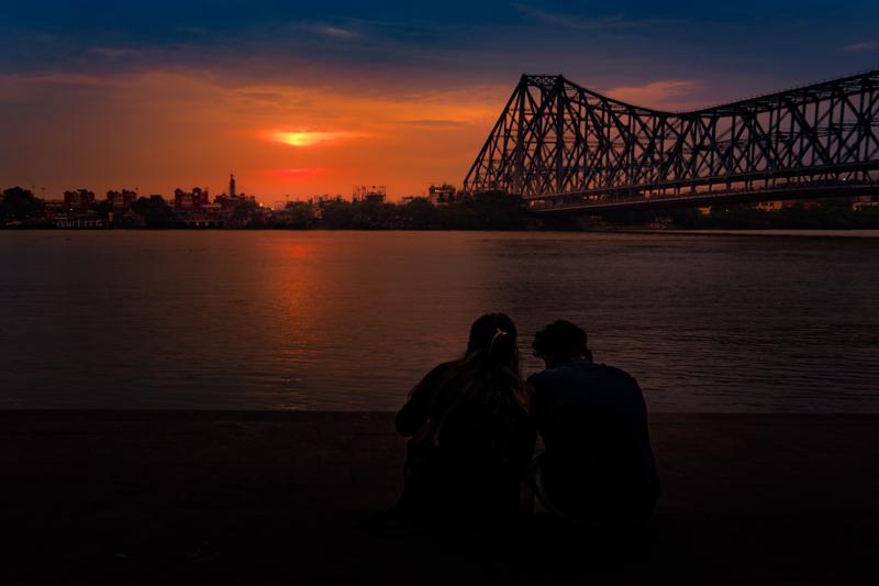 Two figures sit at the bank, facing an orange horizon beneath the bridge; their silhouettes remain small against the wide dark water below.