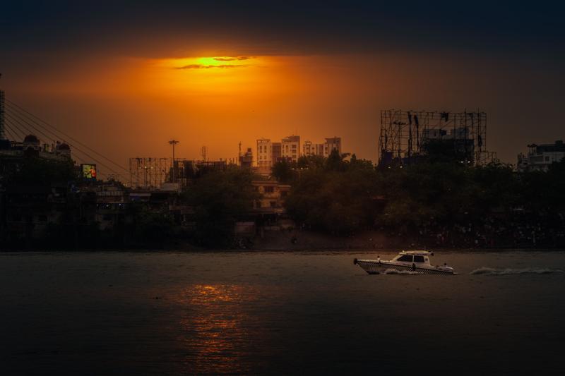The sun sinks over the city as the bridge cuts across the river, its lamps and reflections stretching into the darkening river water.