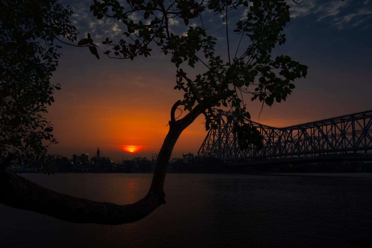 Orange light settles over the river as a tree darkens the foreground, the bridge stretching beyond a lone figure at the water’s edge.