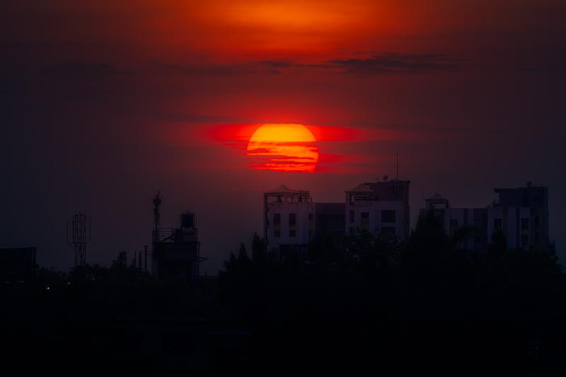 A bright red disc hangs over a low city skyline, its colour fading through smoky orange into the dark foreground, nearby, in silhouette.