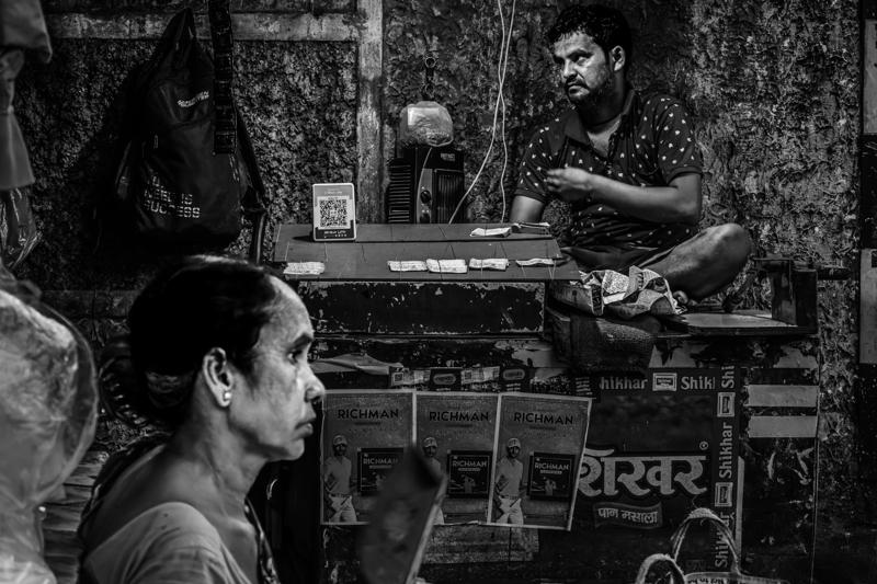 The vendor waits behind his counter as a woman passes in profile; hanging pictures and stacked produce gather around him.