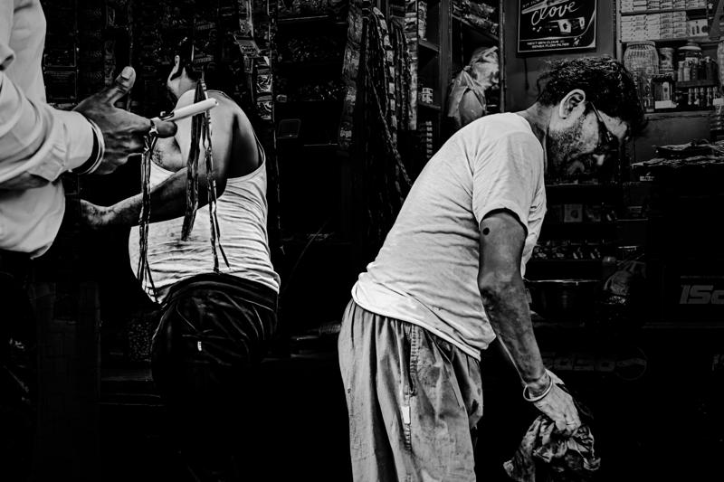 A man leans above a wash tub in the market lane, his clothing reflected in the wet stones around him.